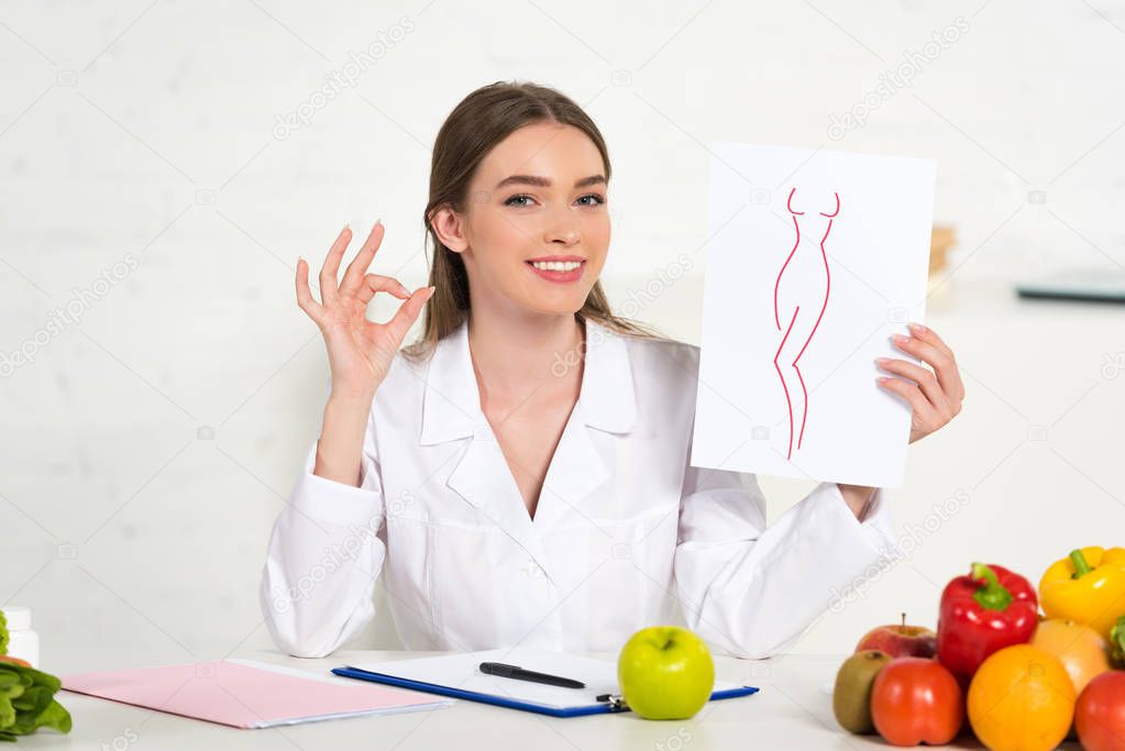 smiling dietitian in white coat holding paper with image of perfect body and showing okay sign at workplace with fruits and vegetables on table