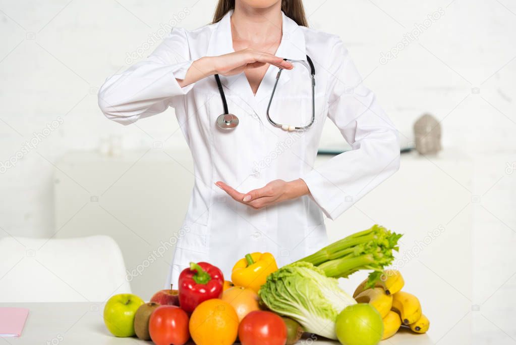 partial view of dietitian in white coat with stethoscope near fresh fruits and vegetables