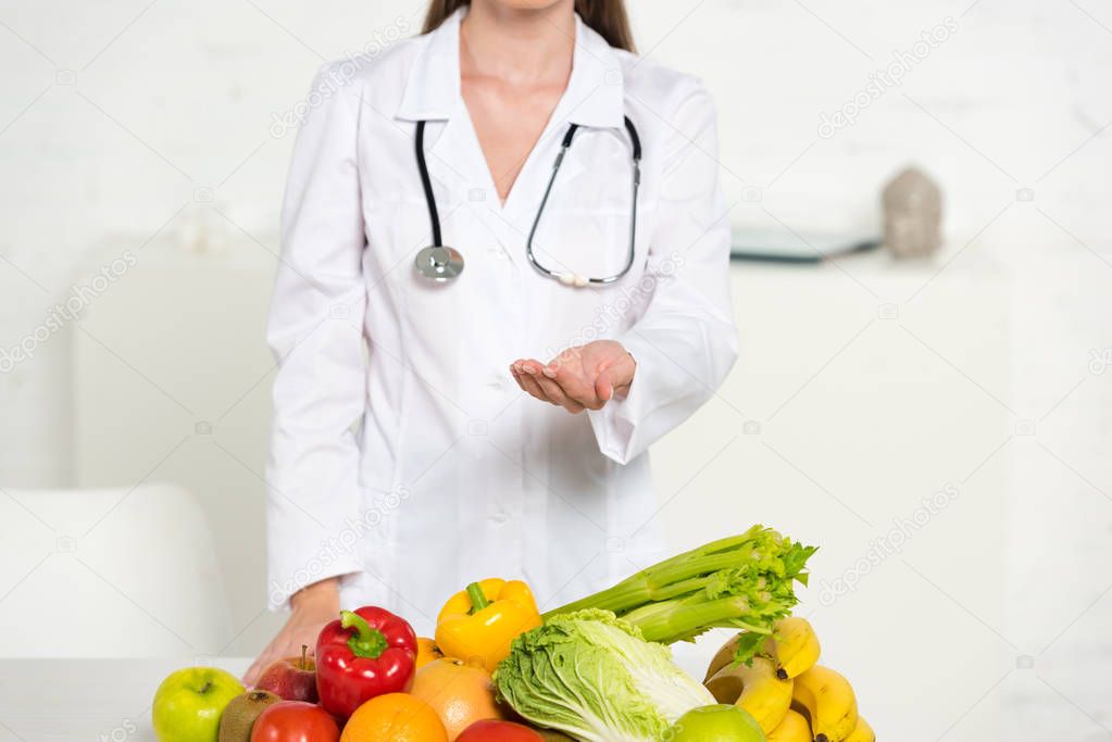cropped view of dietitian in white coat with stethoscope standing with outstretched hand near fresh fruits and vegetables