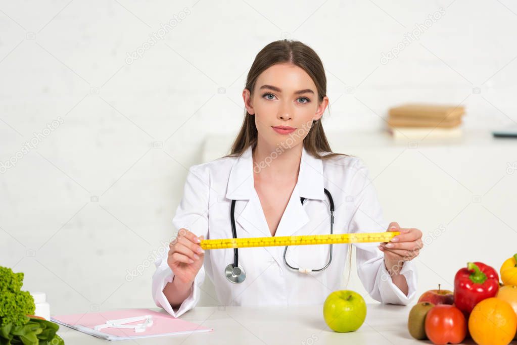 front view of dietitian in white coat with stethoscope holding measure tape at table with fruits and vegetables