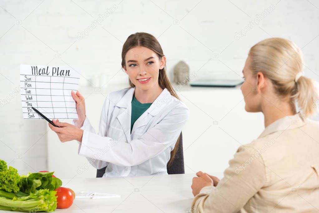 smiling dietitian in white coat holding meal plan and patient at table