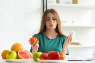 sad woman sitting at table with fruits and vegetables and holding pills and tomato clipart