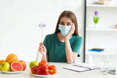 tıbbi maskeli kadın meyve, sebze ve hap ile masada oturan ve evde yazıt alerjisi ile sopa üzerinde afiş tutan