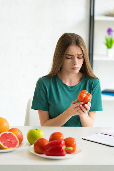 sad woman sitting at table with fruits and vegetables and holding tomato