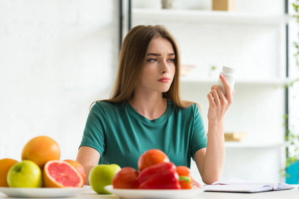 sad woman sitting at table with fruits and vegetables and holding pills