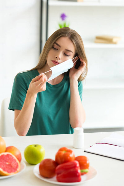 girl in medical mask sitting at table with pills, fruits and vegetables 
