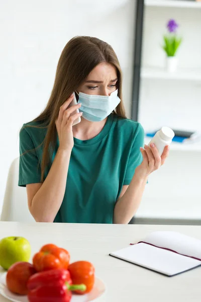 医療マスクを着た女性がスマートフォンで話し 果物や野菜とテーブルで錠剤を見ている — ストック写真