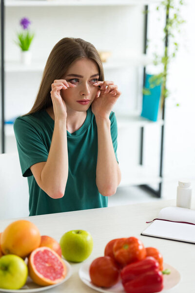 girl with allergy wiping tears while sitting at table with fruits, vegetables and pills