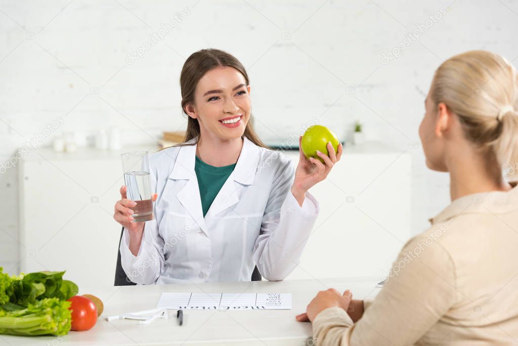 smiling dietitian in white coat holding apple and glass of water and patient at table