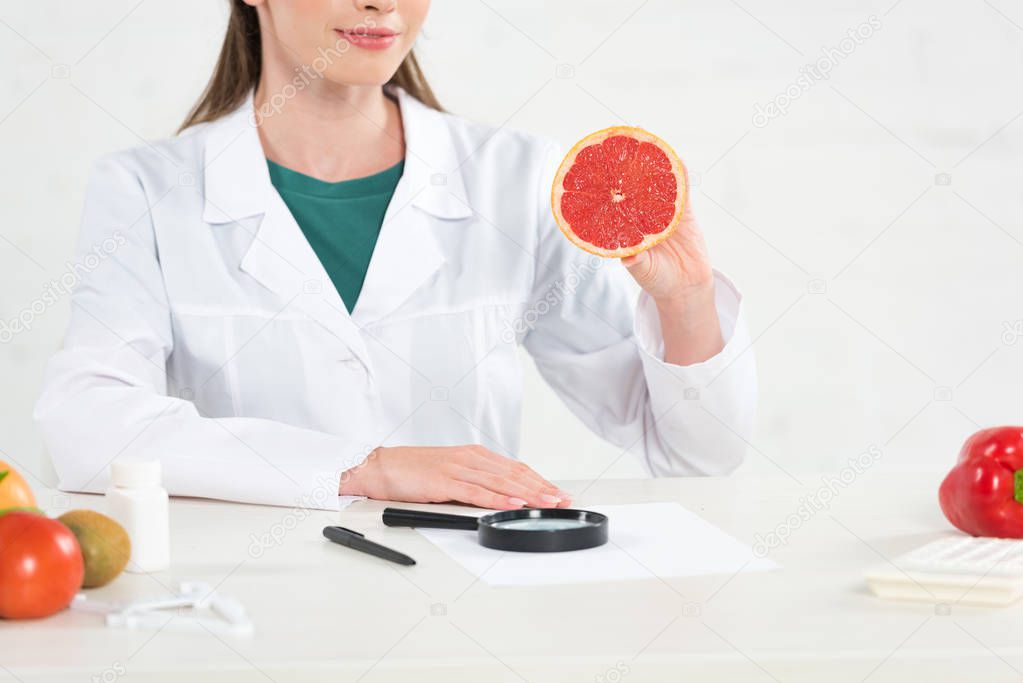 cropped view of dietitian in white coat holding cut grapefruit at workplace