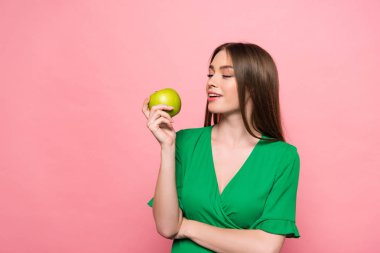 attractive young woman holding green apple and smiling isolated on pink clipart