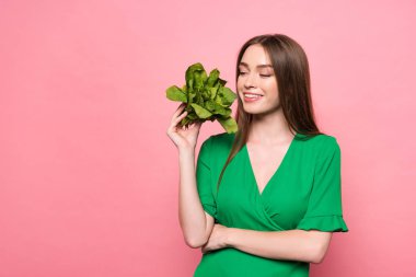 smiling young woman holding spinach with closed eyes isolated on pink