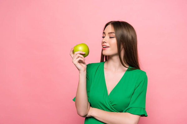 attractive young woman holding green apple and smiling isolated on pink