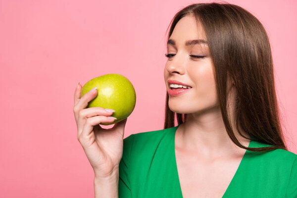 attractive young woman holding green apple and smiling isolated on pink