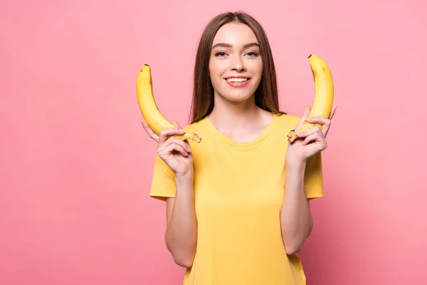 attractive smiling girl holding bananas and looking at camera on pink
