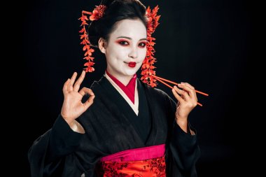 saç çubukları tutan ve siyah izole tamam işareti gösteren siyah kimono güzel geyşa gülümseyen