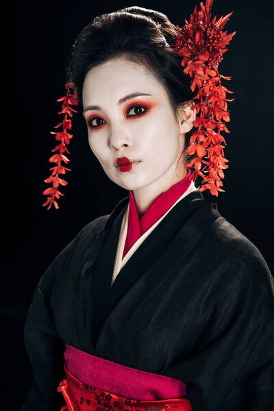 beautiful geisha in black and red kimono and flowers in hair looking at camera isolated on black