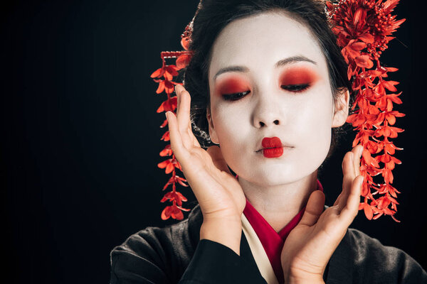 beautiful geisha in black and red kimono and flowers in hair with hands near face looking down isolated on black