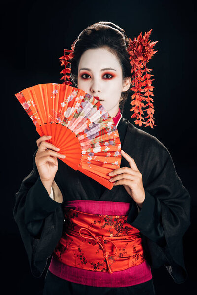 geisha in black kimono with red flowers in hair holding traditional hand fan isolated on black