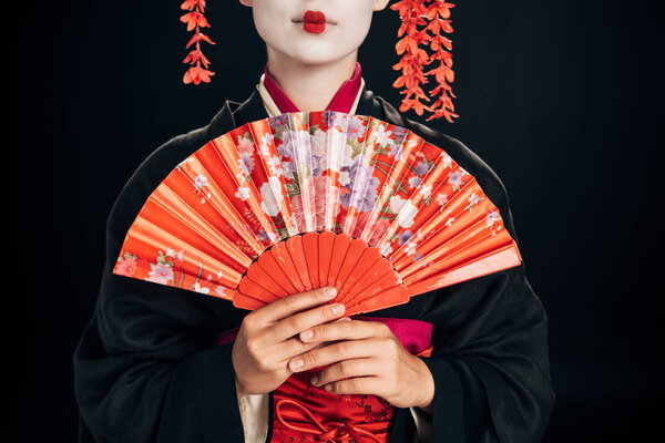 cropped view of beautiful geisha in black kimono with red flowers in hair holding traditional hand fan isolated on black