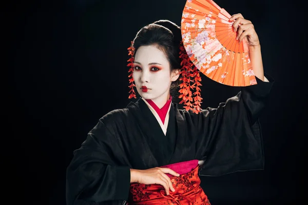 beautiful geisha in black kimono with red flowers in hair posing with traditional hand fan isolated on black
