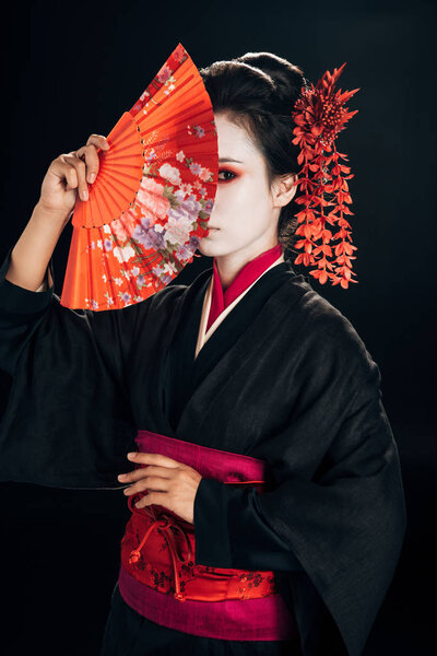 beautiful geisha in black kimono with red flowers in hair hiding behind traditional hand fan isolated on black