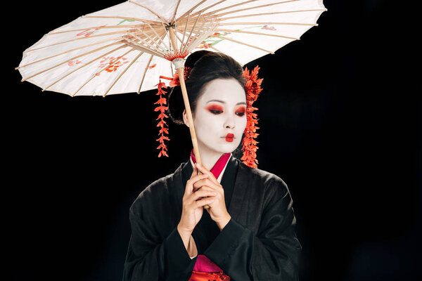 beautiful geisha in black kimono with red flowers in hair holding traditional asian umbrella and looking down isolated on black