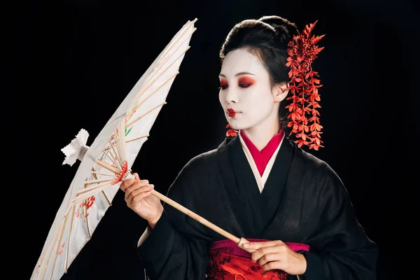 黒い着物の美しい芸者で 黒で孤立した伝統的なアジアの傘を見て髪に赤い花を持つ — ストック写真