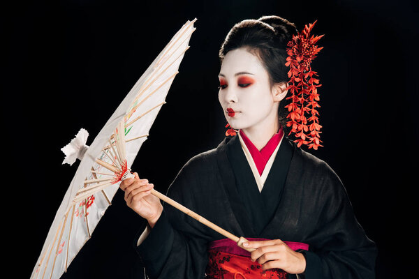 красивая гейша в черном кимоно с красными цветами в волосах, смотрящая на традиционный азиатский зонтик, изолированный на черном
