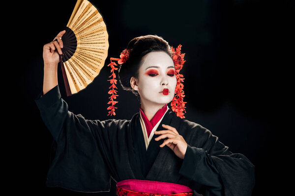 geisha in black kimono with red flowers in hair dancing with traditional asian hand fan isolated on black