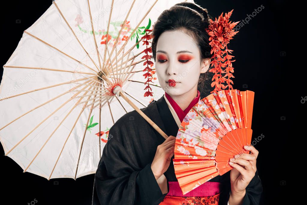 beautiful geisha in black kimono with red flowers in hair holding traditional asian umbrella and hand fan isolated on black