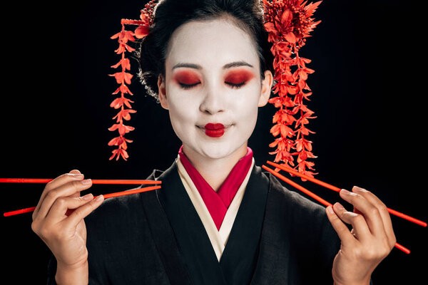 smiling beautiful geisha in black kimono with red flowers in hair and closed eyes holding chopsticks isolated on black