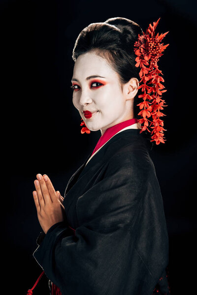 side view of happy beautiful geisha in black and red kimono and flowers in hair looking at camera isolated on black