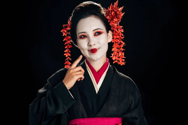 side view of happy beautiful geisha in black and red kimono and flowers in hair pointing with finger at face isolated on black