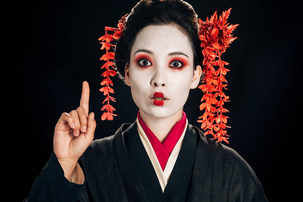 shocked beautiful geisha in black and red kimono and flowers in hair showing idea gesture isolated on black