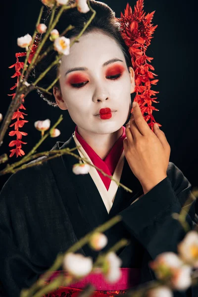 髪に触れる顔に赤い花を着た黒い着物の美しい芸者の選択的な焦点と黒に隔離された桜の枝 — ストック写真