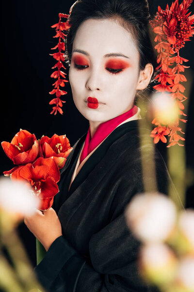 selective focus of beautiful geisha in black kimono with red flowers and sakura branches isolated on black