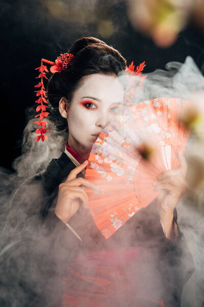 selective focus of beautiful geisha in black kimono with flowers in hair holding hand fan and sakura branches in smoke