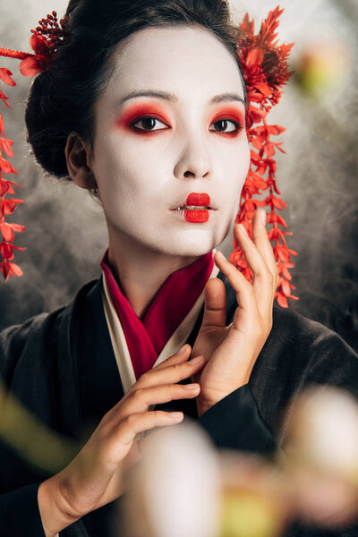 selective focus of beautiful geisha in black kimono with red flowers in hair touching face and sakura branches on black background with smoke