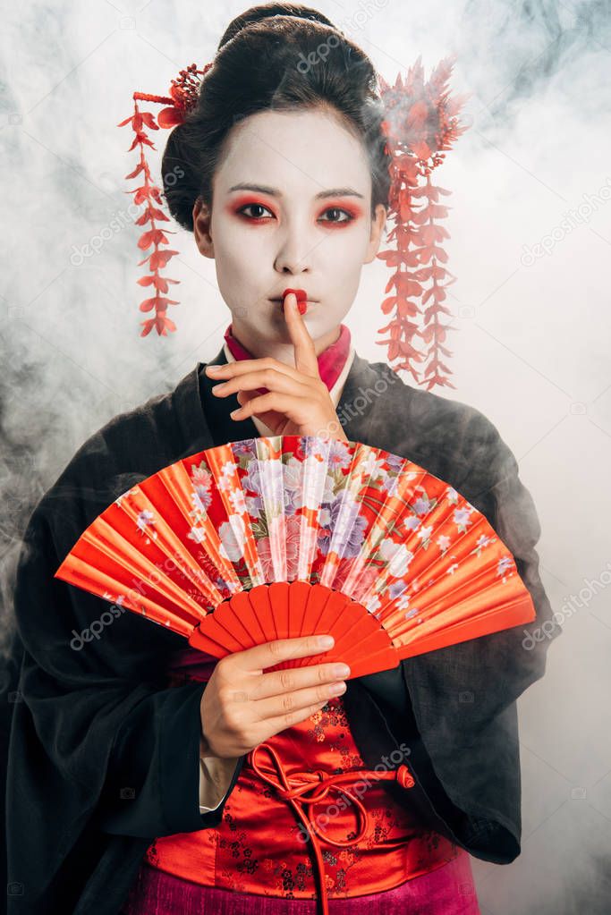 beautiful geisha in black kimono with flowers in hair holding hand fan and showing shh gesture in smoke