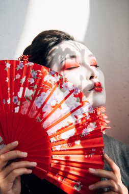 kırmızı ve beyaz makyaj ve kapalı gözleri güneş ışığında el fan tutarak güzel geyşa