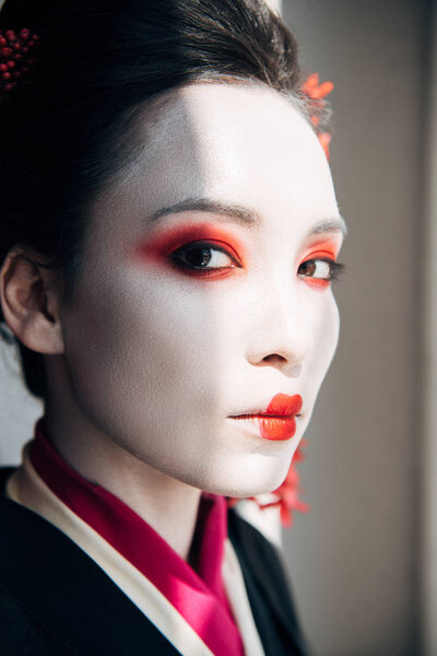 portrait of beautiful geisha with red and white makeup looking at camera in sunlight