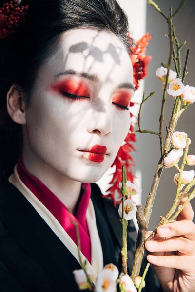 beautiful geisha with red and white makeup and sakura in sunlight