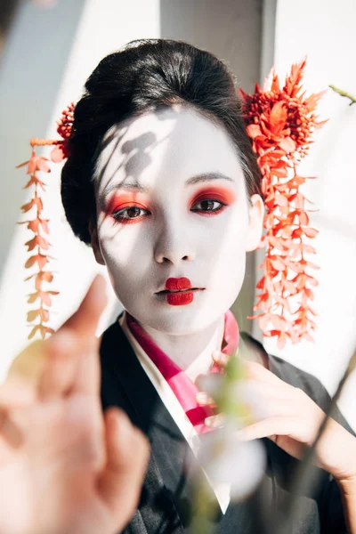 樱花枝和美丽的艺伎与红色和白色的妆容在阳光下手势的选择性焦点 — 图库照片