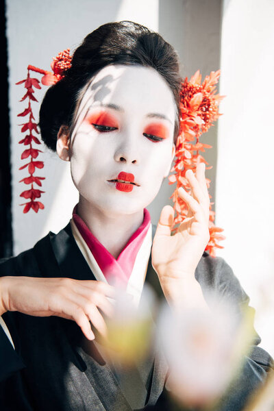 selective focus of sakura branches and beautiful geisha with red and white makeup and closed eyes in sunlight