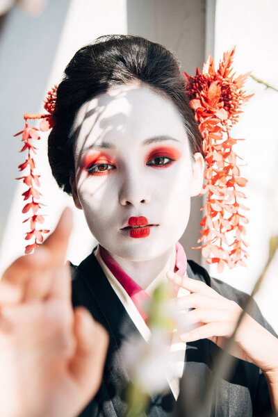 selective focus of sakura branches and beautiful geisha with red and white makeup in sunlight