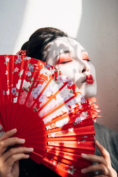 beautiful geisha with red and white makeup and closed eyes holding hand fan in sunlight