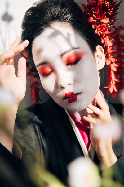 selective focus of tree branches and geisha with red and white makeup and closed eyes in sunlight
