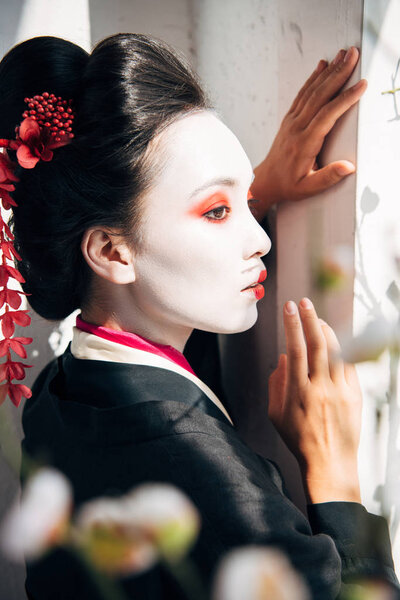 selective focus of sakura branches and beautiful geisha near white wall in sunlight