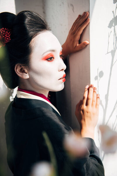 selective focus of sakura branches and beautiful geisha near wall in sunshine